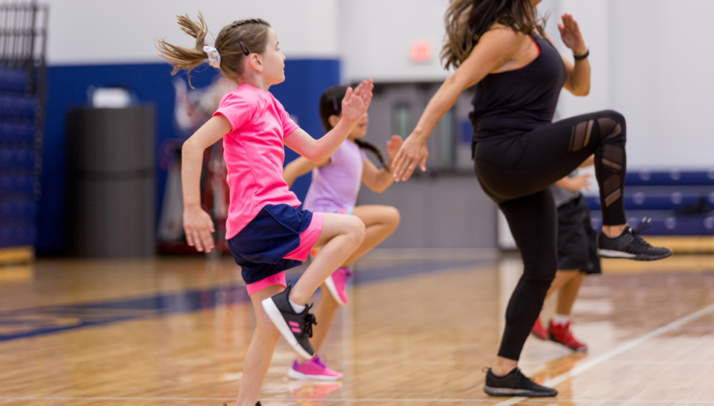 Picture of a school fitness class where a child is standing on one knee behind a fitness instructor