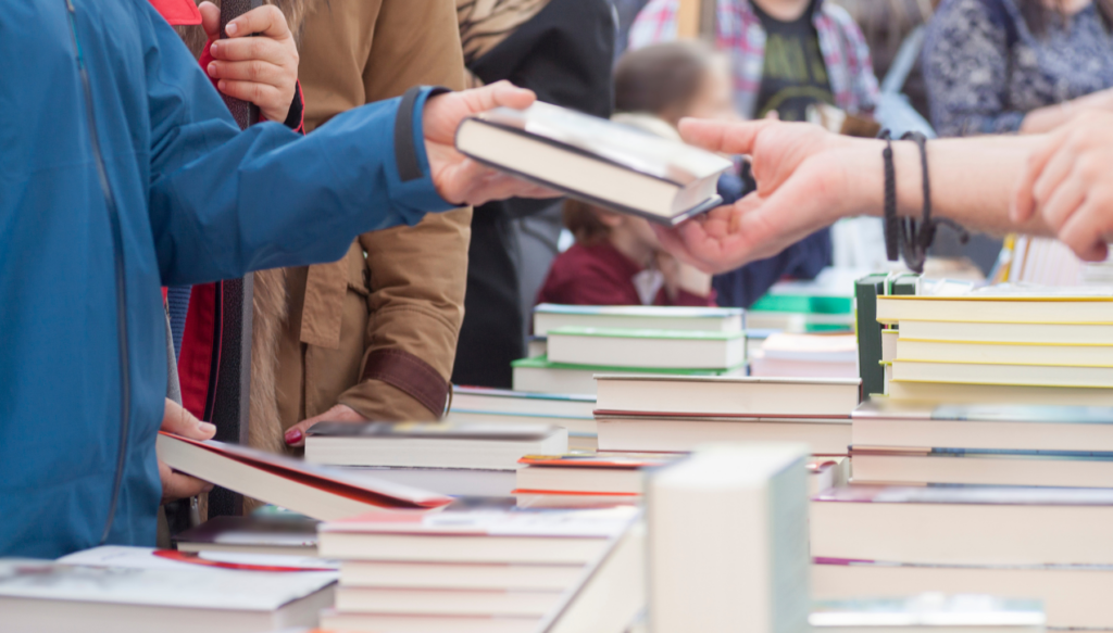 picture of someone handing another person two books. There is a table of books underneath