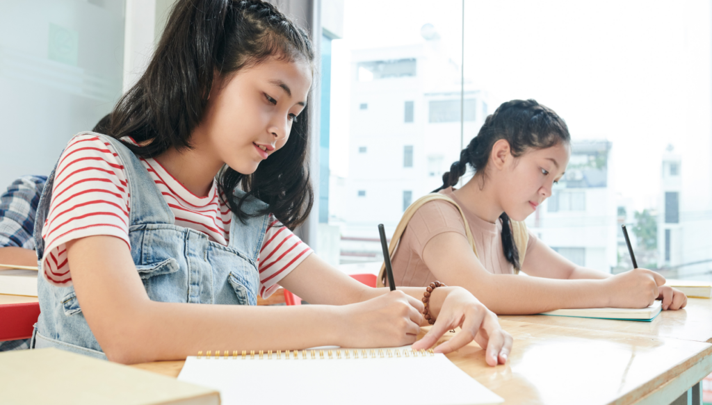 picture of two students writing in a notebook at a desk in a classroom